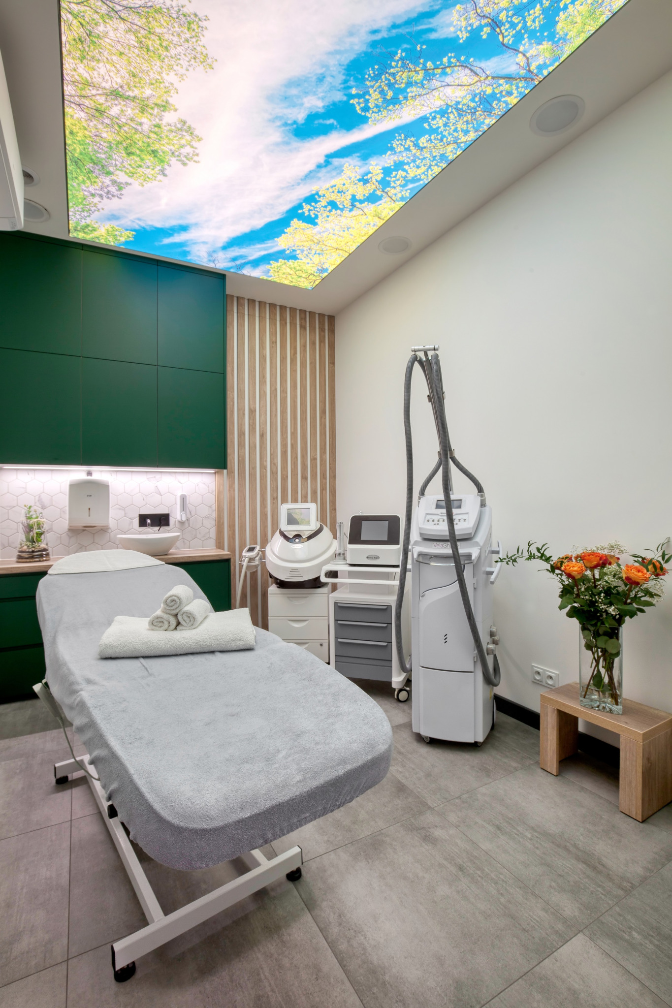 Jasne wnętrze gabinetu kosmetycznego z łóżkiem do zabiegów, nowoczesną aparaturą, bukietem różowych róż i podświetlanym sufitem imitującym niebo z drzewami.