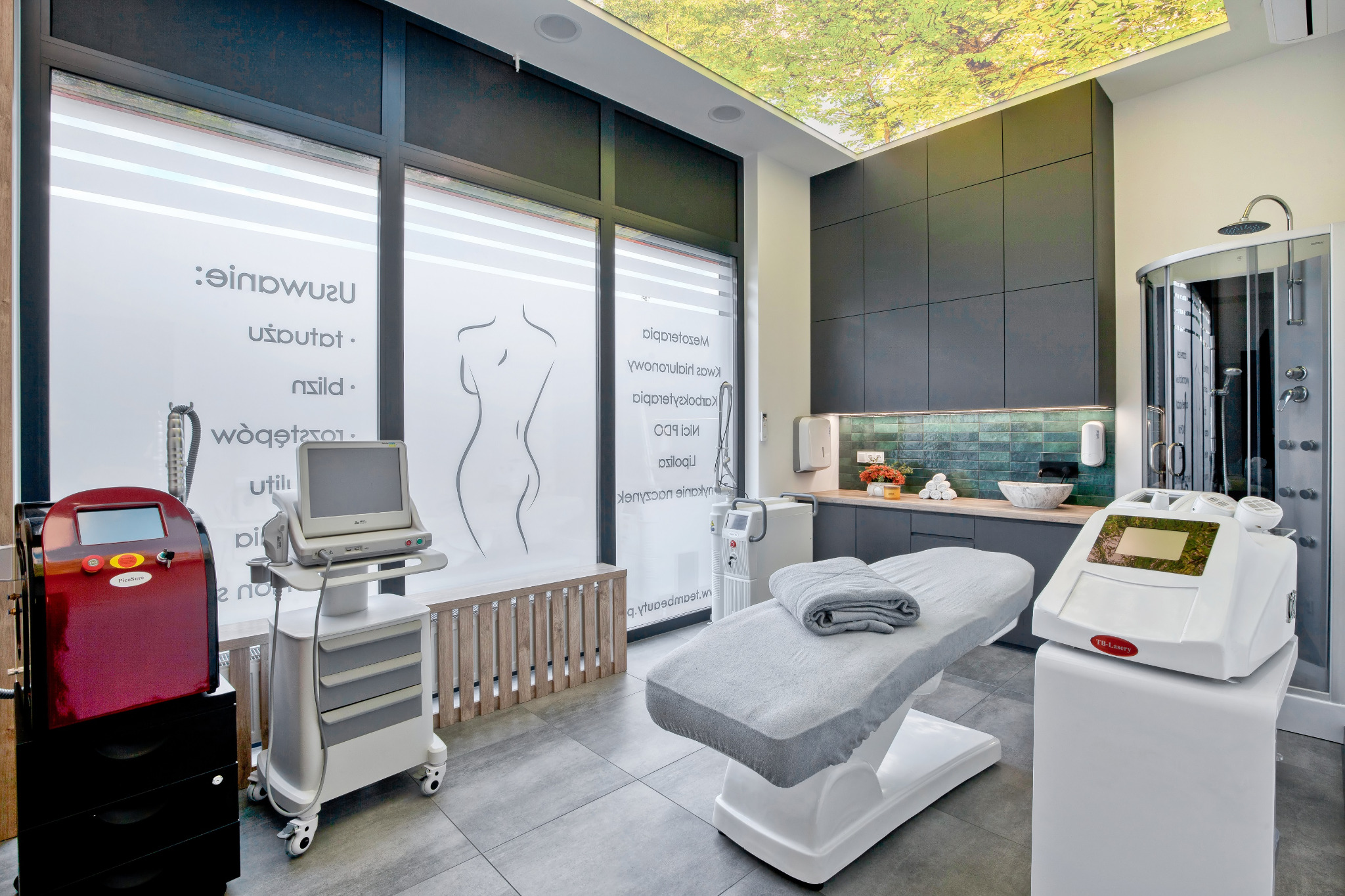 Nowoczesny gabinet kosmetyczny z fototapetą drzew na suficie, wyposażony w profesjonalne urządzenia do zabiegów na ciało, leżankę z szarym ręcznikiem i kabinę prysznicową.