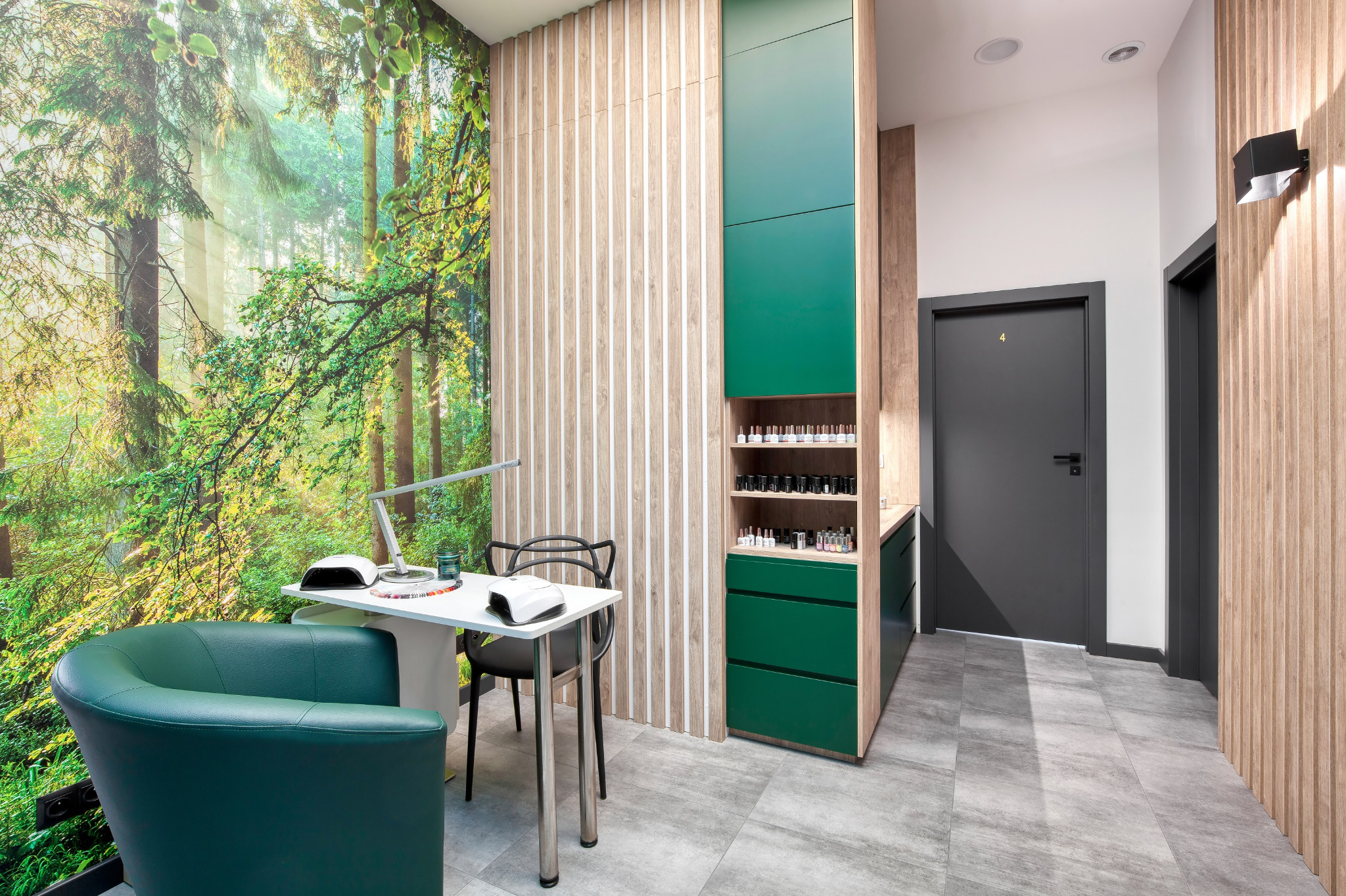 Wnętrze salonu kosmetycznego z fototapetą leśną, stanowiskiem do manicure z lampą UV i zielonym fotelem oraz szafką z lakierami do paznokci w odcieniach zieleni i brązu.