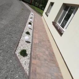 Ułożona kostka brukowa w odcieniach brązu tworząca ścieżkę obok domu, z ozdobnym pasem białych kamieni, kulistych lamp i krzewów.