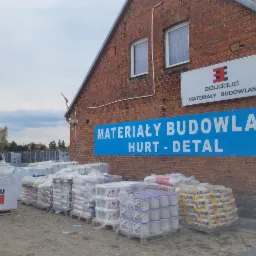 Składowane materiały budowlane przed budynkiem z szyldem informującym o sprzedaży hurtowej i detalicznej, widoczne palety z workami i farbami.