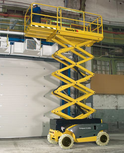 Żółto-czarny podnośnik nożycowy Haulotte Compact 12 z operatorem w niebieskim kombinezonie i białym kasku, ustawiony w hali przemysłowej o szarych ścianach i widocznych instalacjach.