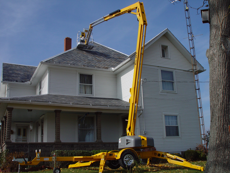Żółty podnośnik na przyczepie ustawiony przy białym, dwupiętrowym domu z szarym dachem, osoba w koszu podnośnika pracuje przy kominie.