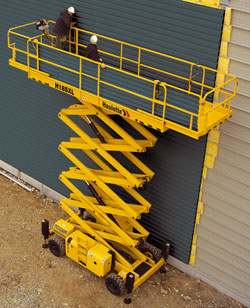 Żółty podest roboczy nożycowy marki Haulotte M165XL z dwoma pracownikami w kaskach, na tle zielonej blachy trapezowej i fragmentu jasnej ściany budynku.