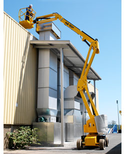 Żółty, przegubowo-teleskopowy podest ruchomy HA 15 IP podczas pracy na zewnątrz budynku przemysłowego, operator w koszu zabezpieczony kaskiem, widoczny system wentylacyjny na elewacji budynku.
