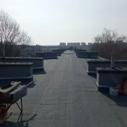 Widok z góry na płaski dach pokryty papą termozgrzewalną z kominami wentylacyjnymi, w tle zabudowa miejska i drzewa bez liści, wózek z rolkami papy i butla gazowa z narzędziami dekarskimi.