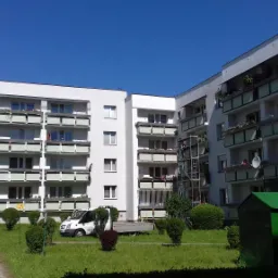 Widok na ocieplony budynek wielorodzinny z balkonami, na trawniku zaparkowany biały bus dostawczy, obok zielony kontener.