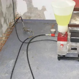 Sprzęt do iniekcji w ścianę z widocznymi dyszami iniekcyjnymi w murze, żółta ciecz w zbiorniku, manometr i przewody.