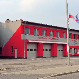 Budynek Ochotniczej Straży Pożarnej i Gminnej Biblioteki Publicznej w Starej Błotnicy, pomalowany w szaro-czerwone pasy, z trzema bramami garażowymi i masztami flagowymi.