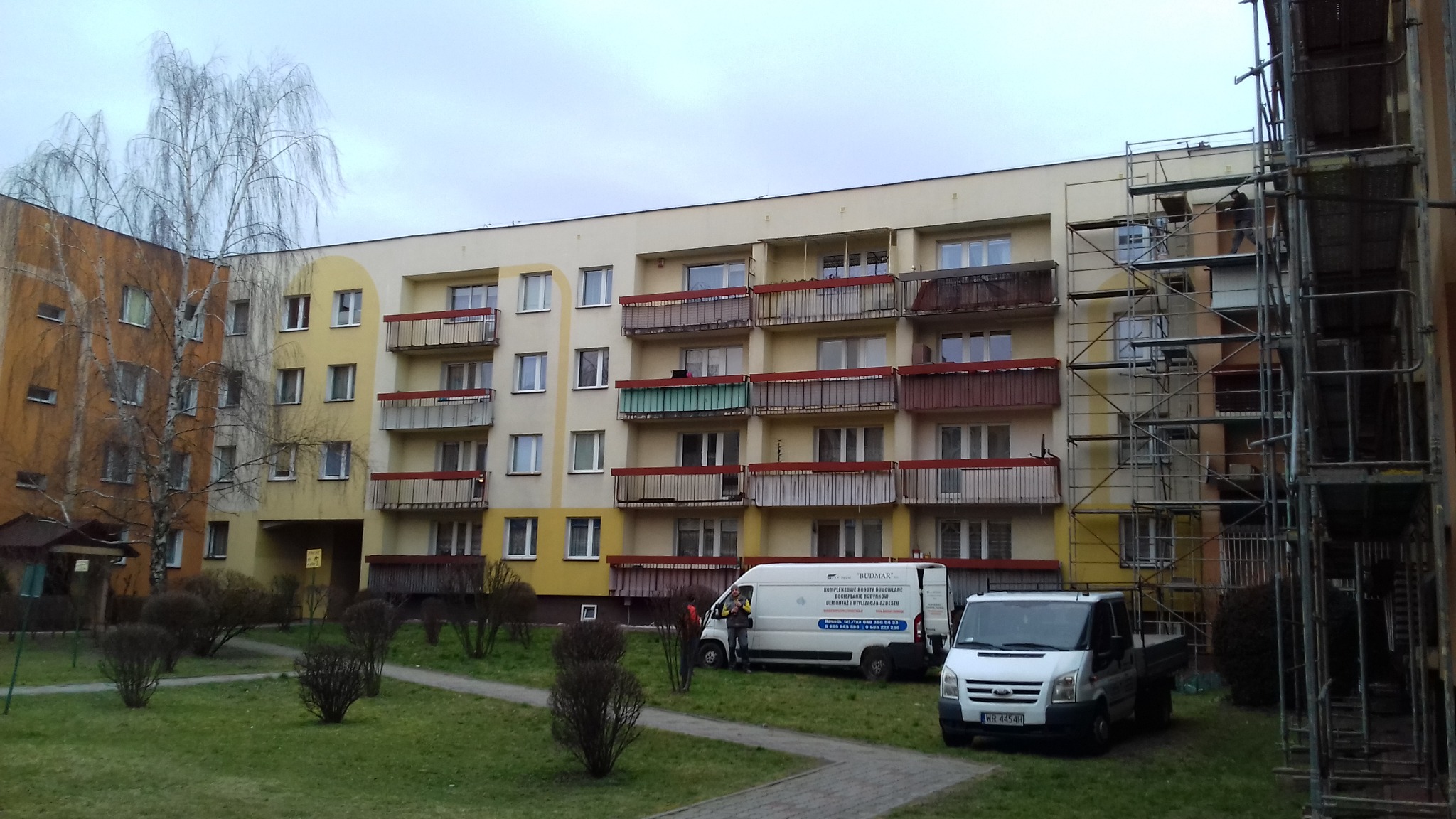Wielopiętrowy blok z balkonami w trakcie prac elewacyjnych, widoczne rusztowania, samochody dostawcze firmy budowlanej na trawniku przed budynkiem, pochmurne niebo.