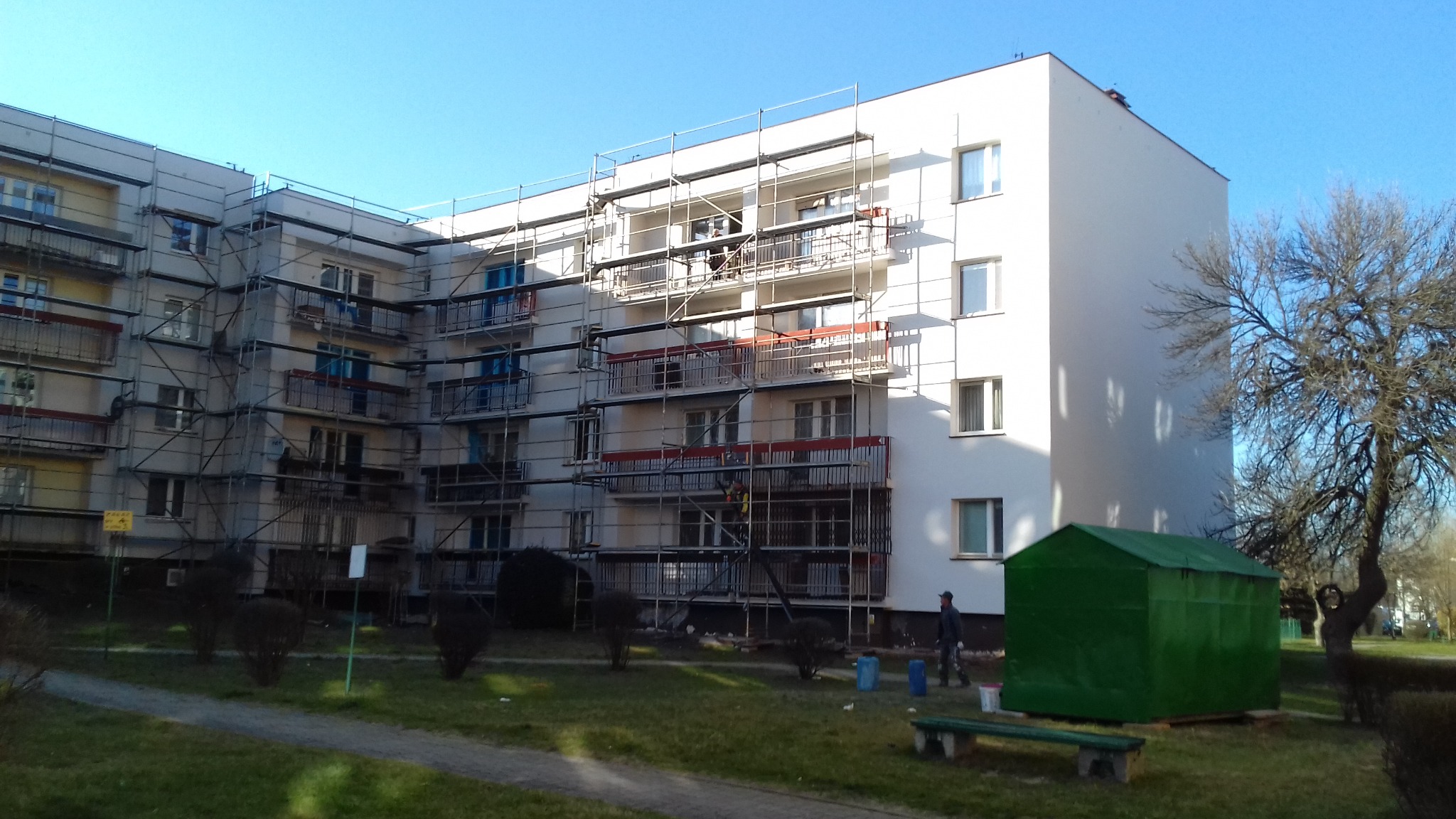 Wielopiętrowy budynek w trakcie prac, otoczony rusztowaniami, z widocznymi pracownikami na rusztowaniach, świeżo pomalowana ściana szczytowa, na pierwszym planie zielony blaszak, wiaderka z farbą...