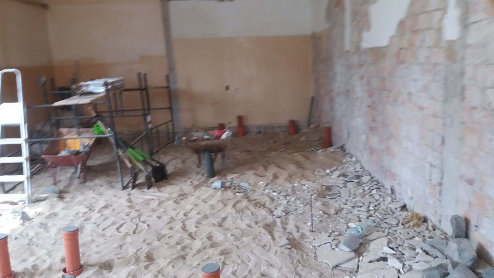 Wnętrze w trakcie remontu: gruz, piasek, rusztowania, drabina i narzędzia w nieuporządkowanym pomieszczeniu z częściowo usuniętym tynkiem na ścianach.