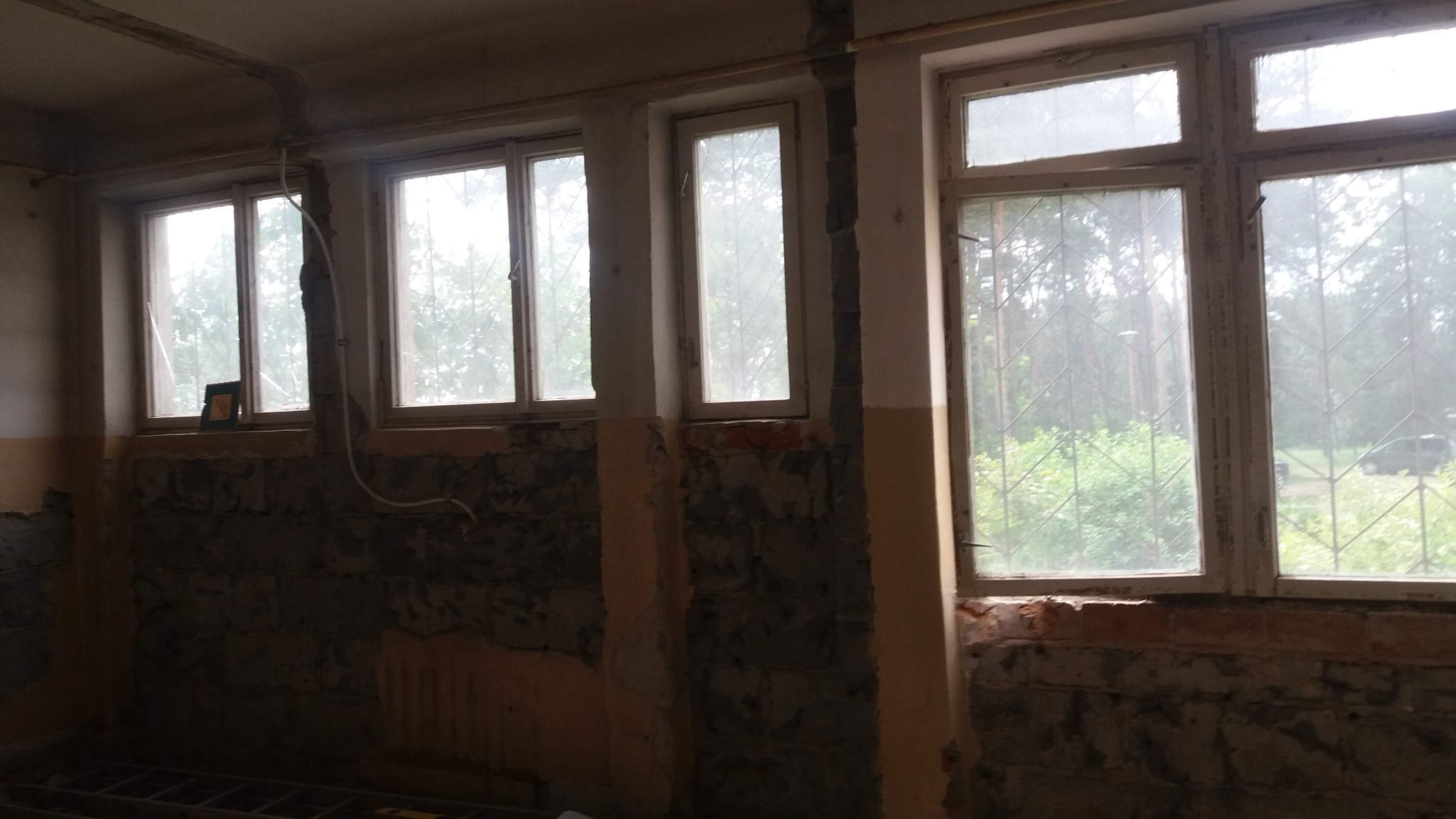 Wnętrze pokoju w trakcie remontu: stare okna z kratami, ściany z odsłoniętą cegłą i resztkami tynku, widoczne rury i instalacje elektryczne.