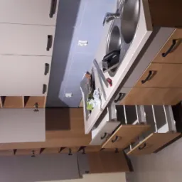 Kuchnia z drewnianymi szafkami i szarymi frontami, widoczne otwarte szuflady i elementy montażowe pod szafkami wiszącymi.