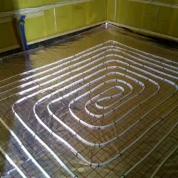 Rozłożona instalacja ogrzewania podłogowego na folii izolacyjnej z siatką, widoczne rury w kształcie meandrów, żółte ściany w tle.