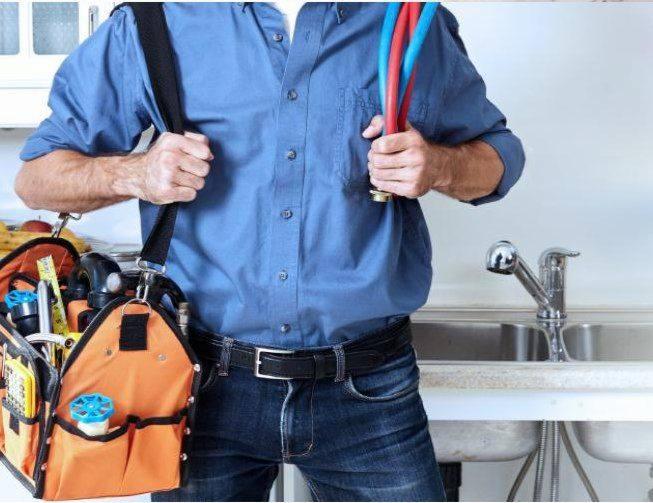 Mężczyzna w niebieskiej koszuli z torbą narzędziową na ramieniu i przewodami w ręku, stojący przed zlewem.
