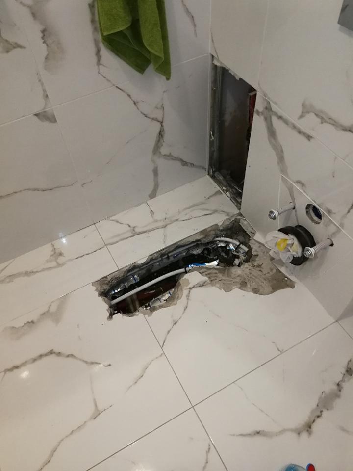 Otwór w podłodze łazienki z widocznymi rurami instalacji wodnej i kanalizacyjnej, ślady po kuciu płytek, fragment ściany z otworem rewizyjnym, ręcznik na ścianie.