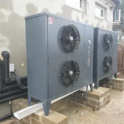 Dwie szare jednostki zewnętrzne z wentylatorami, marki Feris, zamontowane na betonowych podstawach przy ścianie budynku z rurami w izolacji.