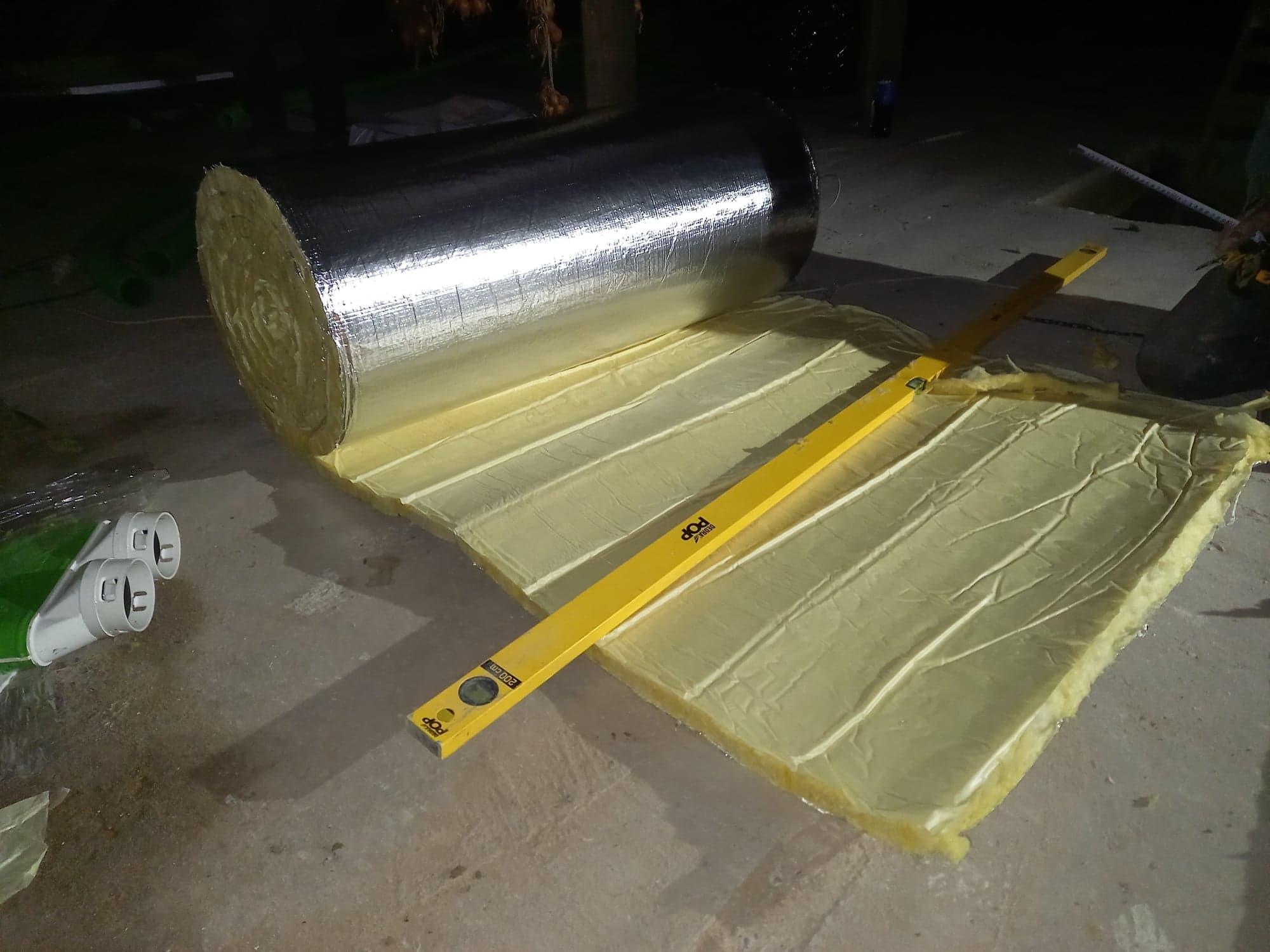 Rolka izolacji z folią aluminiową rozwijana na podłodze, żółta poziomica leży na izolacji, widoczne rury wentylacyjne i fragment nogi montera.