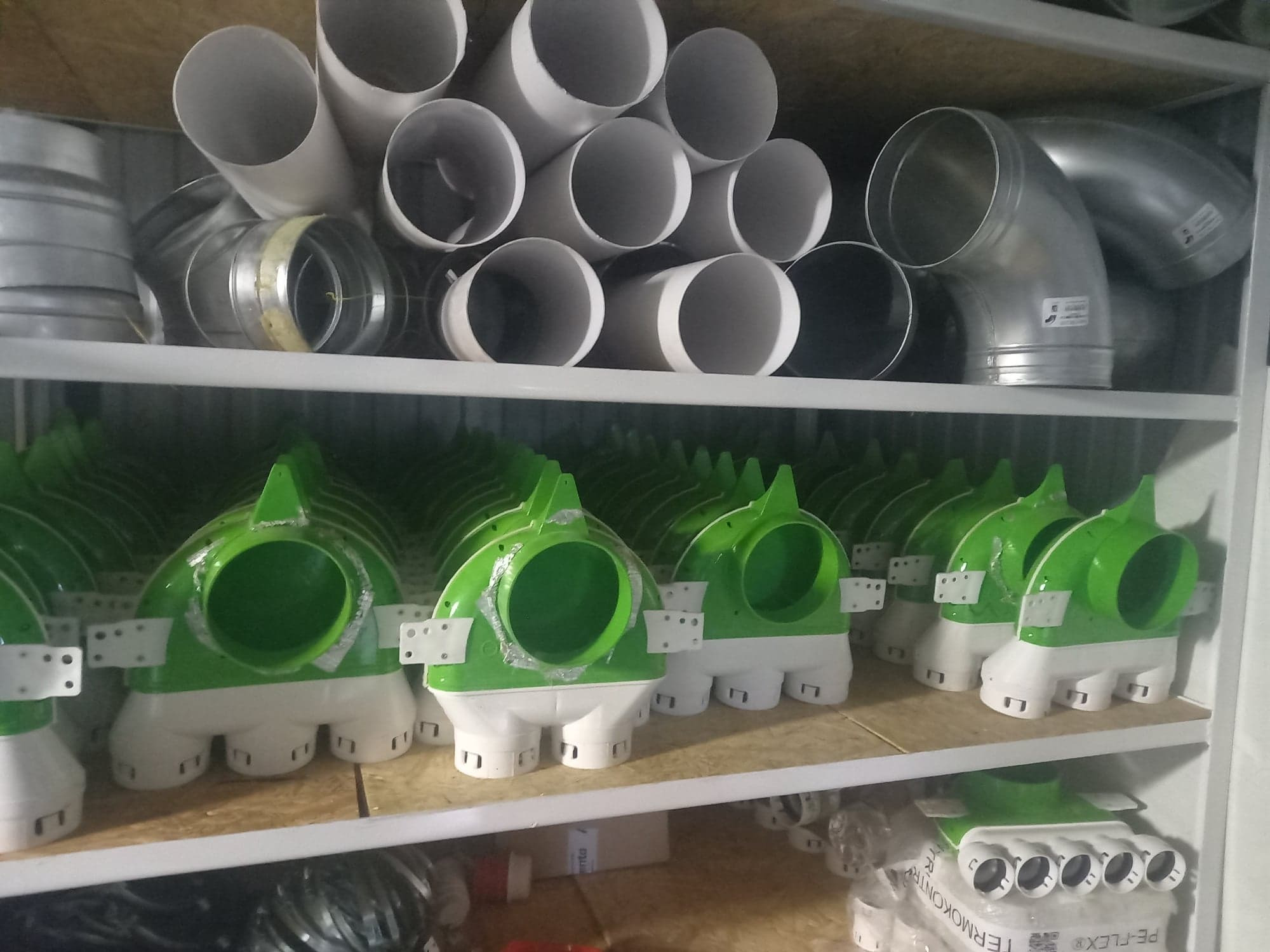Półki magazynowe wypełnione elementami instalacji wentylacyjnych: rury PCV, kształtki rozdzielcze z zielonymi elementami i białą podstawą. Widoczne również kolanka metalowe.