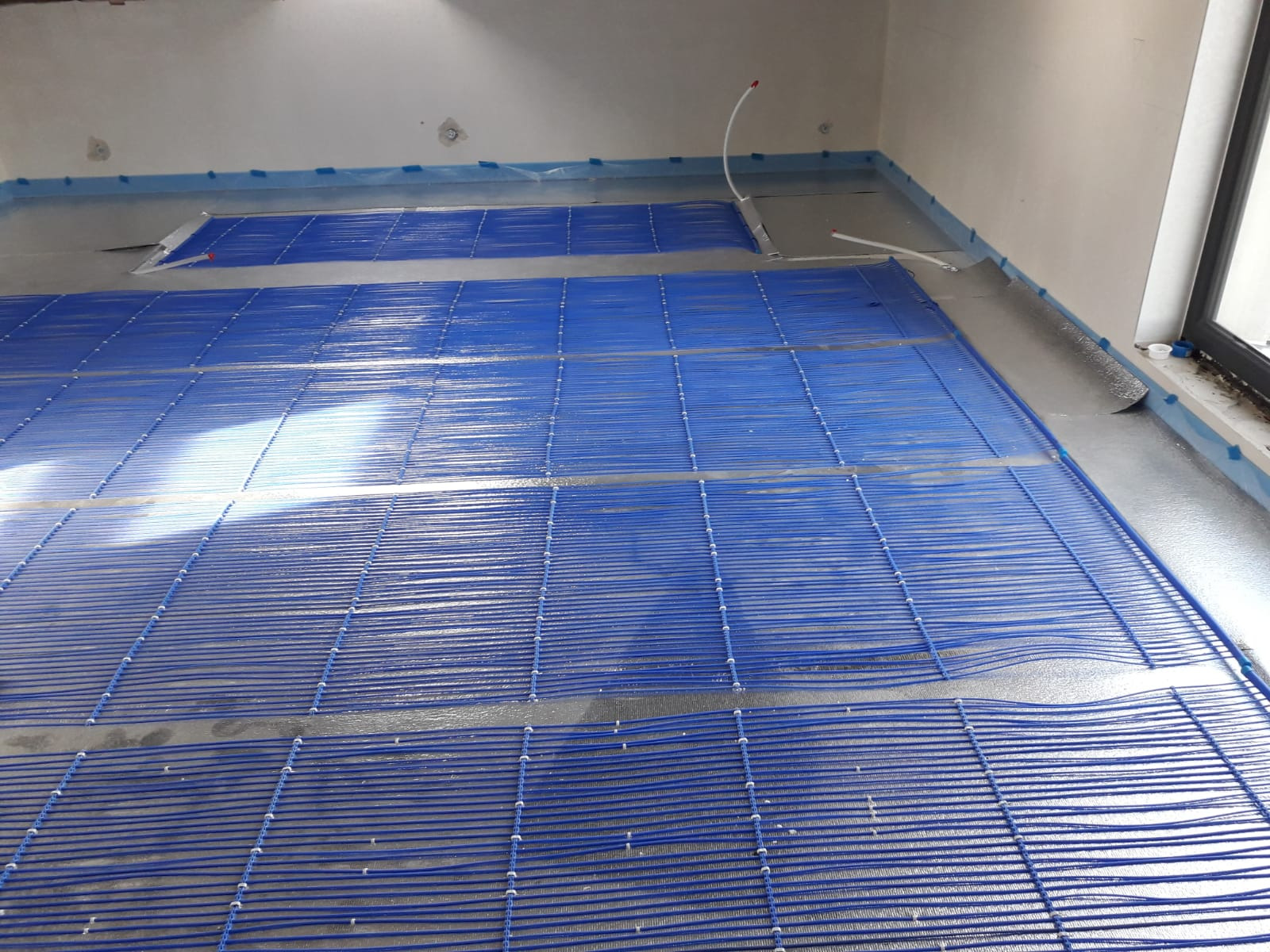 Niebieska mata grzewcza podłogowa rozłożona na szarym podłożu w jasnym pomieszczeniu, widoczne rurki doprowadzające i folia izolacyjna przy ścianach.