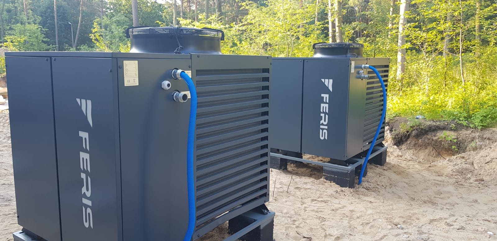 Dwie szare pompy ciepła marki FERIS ustawione na paletach na piaszczystym podłożu z widocznymi niebieskimi rurami przyłączeniowymi i zielonym lasem w tle.