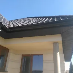 Nowoczesny dom jednorodzinny z ciemną dachówką ceramiczną i czarnymi rynnami, widok z dołu na fragment dachu i elewacji.