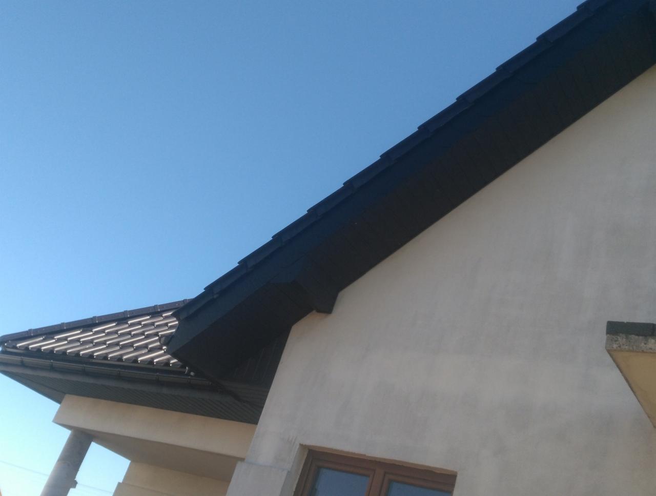 Fragment domu z nowym, ciemnym pokryciem dachowym i jasną elewacją, widoczne okno i błękitne niebo w tle.