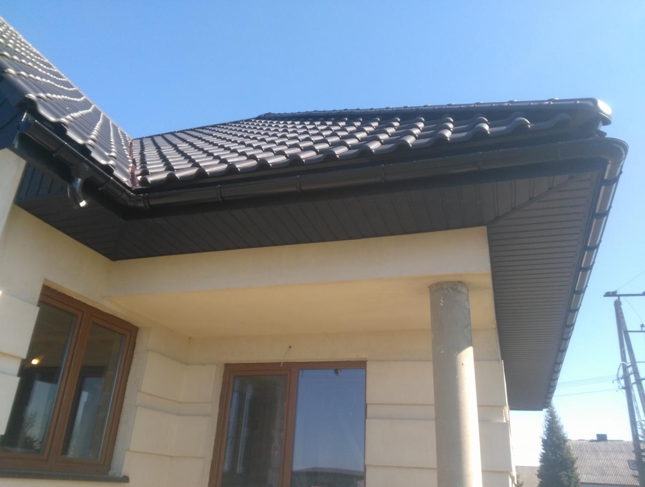 Nowoczesny dom jednorodzinny z ciemną dachówką ceramiczną i czarnymi rynnami, widok z dołu na fragment dachu i elewacji.
