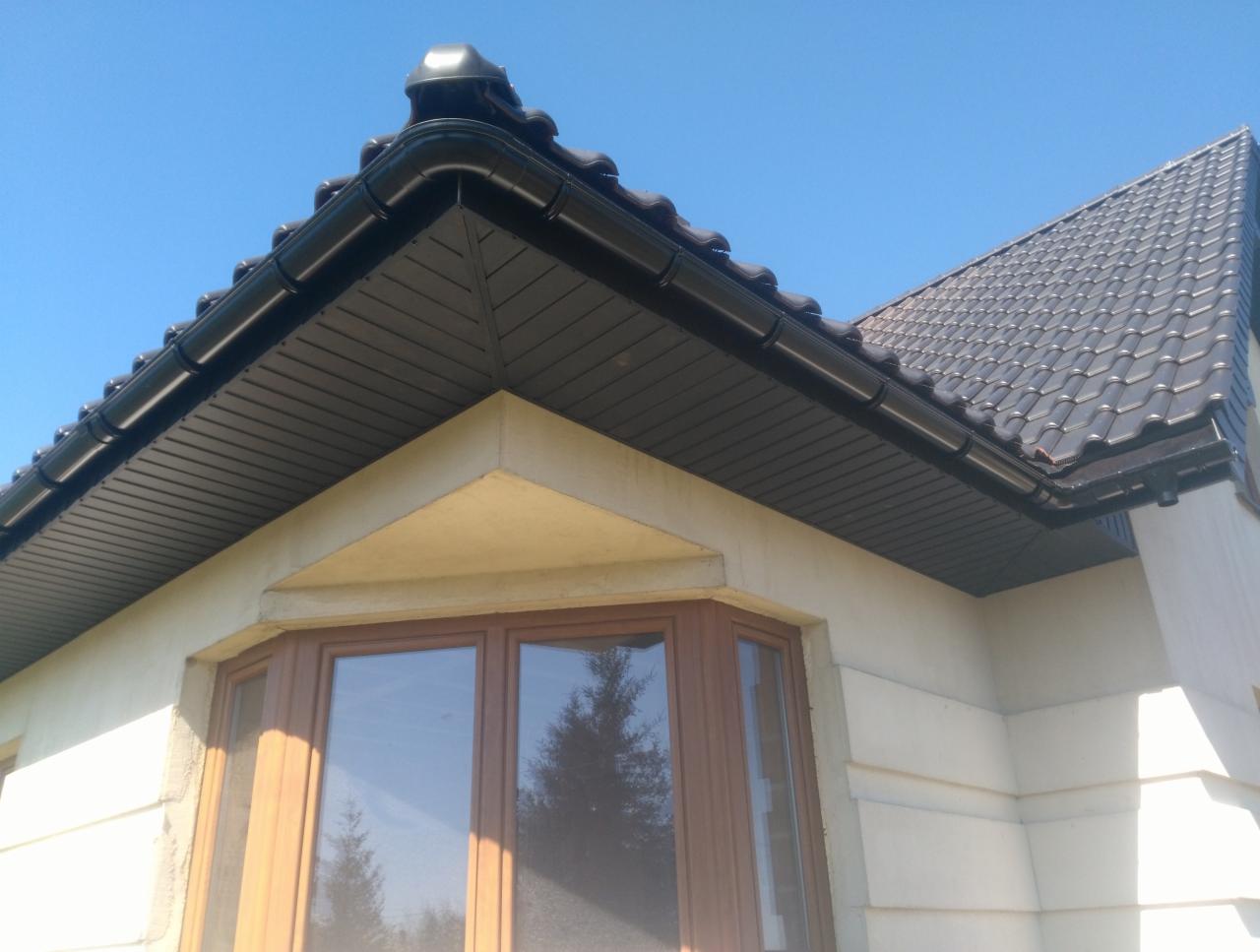 Fragment brązowego dachu z czarnymi rynnami i podbitką, widoczny z dołu na tle błękitnego nieba i okna wykuszowego.
