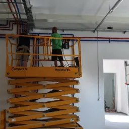 Dwóch mężczyzn na podnośniku nożycowym Haulotte Compact 14 instaluje rury w kolorach czerwonym i niebieskim pod sufitem w nowym, jasnym pomieszczeniu z widoczną bramą garażową w tle.