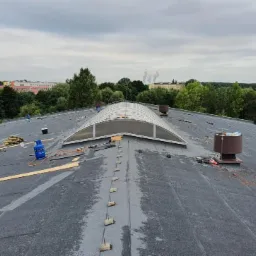 Czarny, płaski dach pokryty papą z centralnie umieszczonym świetlikiem łukowym i widocznymi elementami mocującymi, w tle zieleń drzew i budynki.