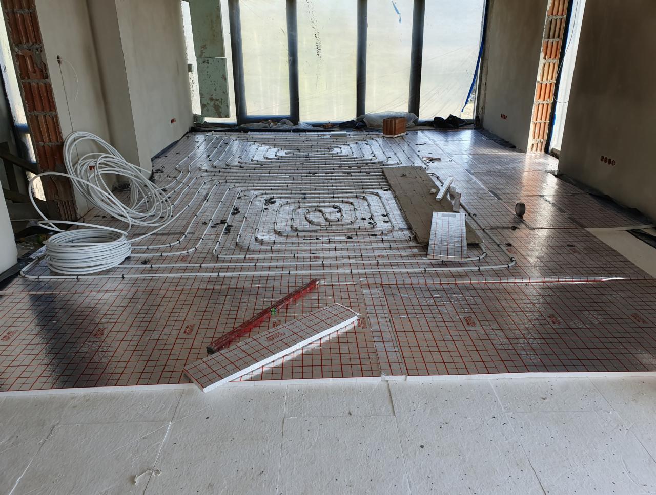 Układanie instalacji ogrzewania podłogowego na folii z siatką, widoczne rury, narzędzia i surowy stan pomieszczenia z oknami.