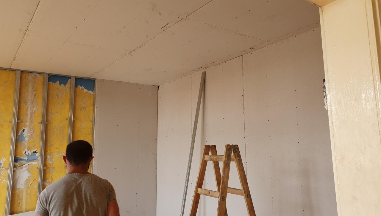 Wnętrze w trakcie remontu: widoczne płyty gipsowo-kartonowe na ścianach i suficie, fragment metalowego stelażu, drewniana drabina i pracownik w szarej koszulce