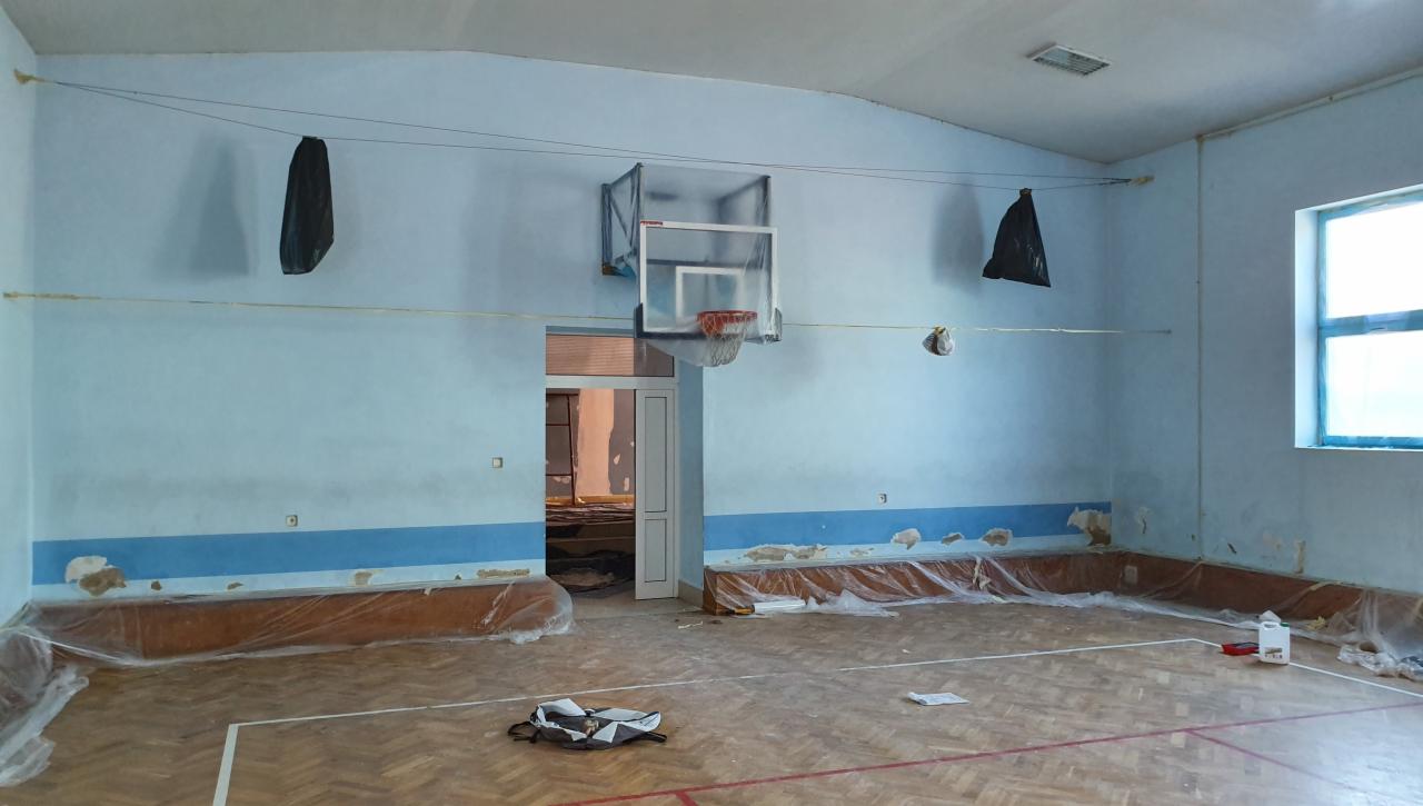Sala gimnastyczna w Kaliszu przygotowywana do malowania: ściany z częściowo usuniętą farbą, zabezpieczone folią dolne partie ścian, kosz do koszykówki, okno z zaklejoną szybą, worki foliowe...