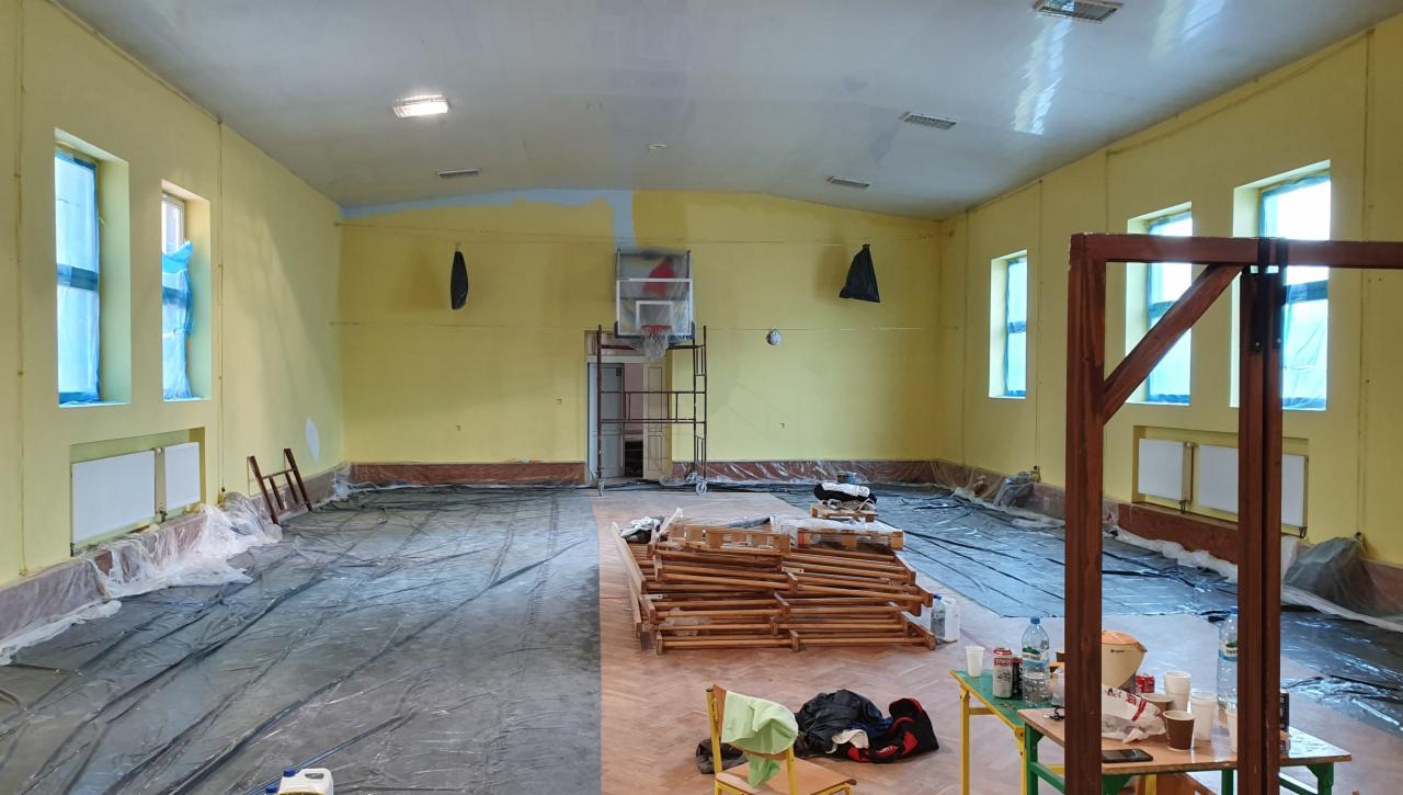 Sala gimnastyczna w trakcie odświeżania: żółte ściany, zabezpieczona podłoga folią, fragment ściany z naniesioną jasnoniebieską farbą, drabina przy koszu, widoczne grzejniki i okna zabezpieczone...