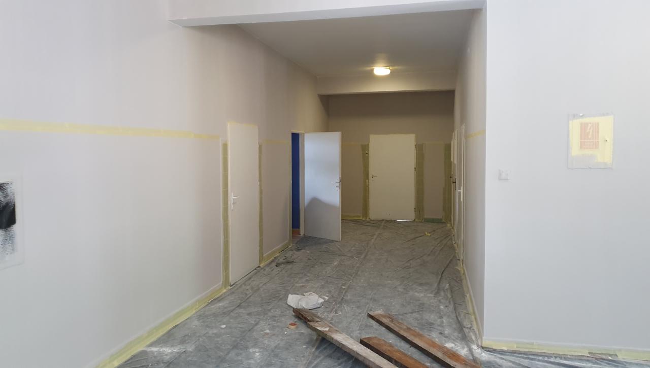 Korytarz w trakcie odświeżania, ściany zabezpieczone taśmą malarską, podłoga przykryta folią ochronną, widoczne deski i resztki materiałów budowlanych.