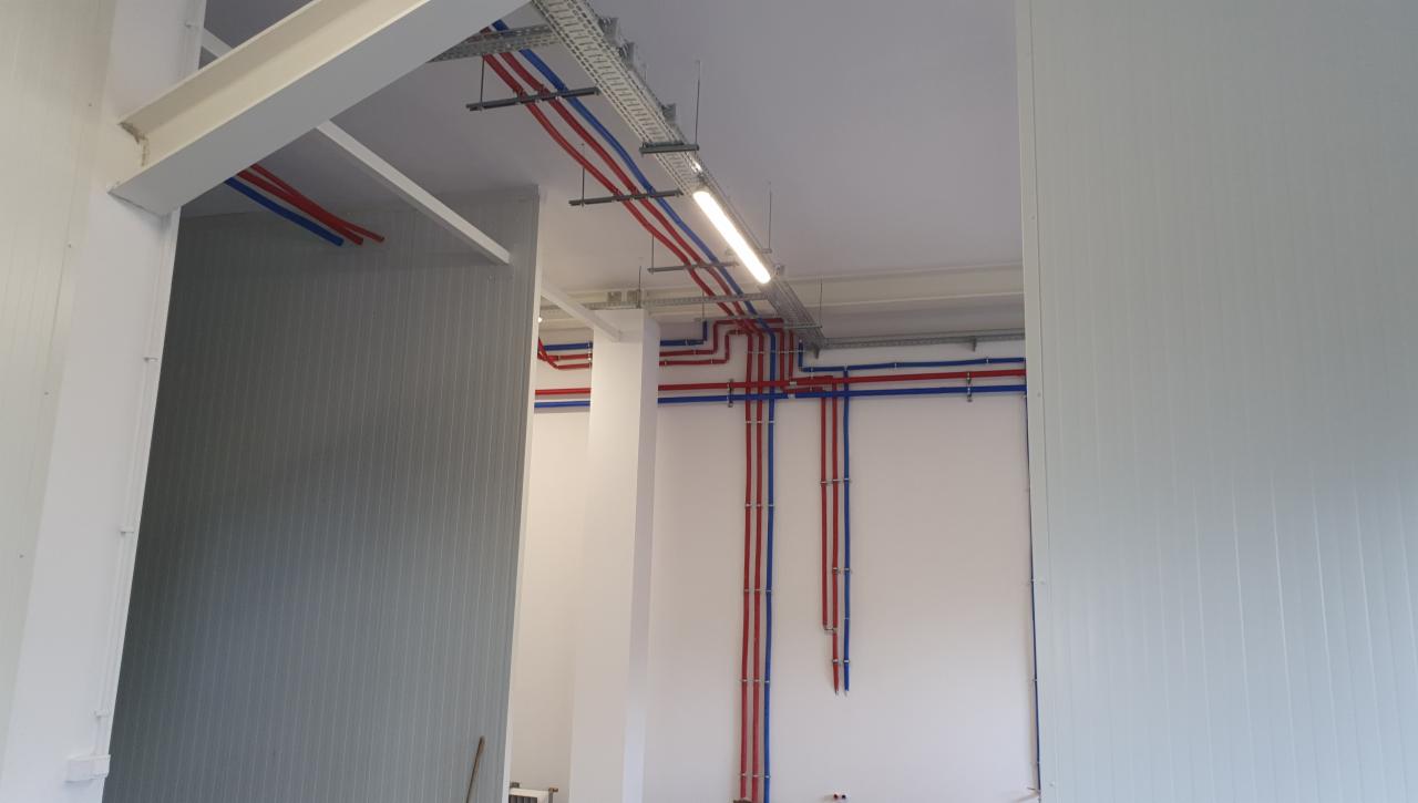 Wnętrze hali produkcyjnej z widocznymi instalacjami hydraulicznymi w kolorze czerwonym i niebieskim, poprowadzonymi wzdłuż białych ścian i pod sufitem, oświetlonymi lampą jarzeniową.