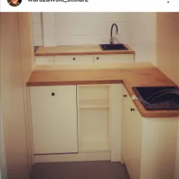 Niewielka kuchnia z białymi szafkami i drewnianym blatem, czarny zlew w rogu, widok z niskiej perspektywy.