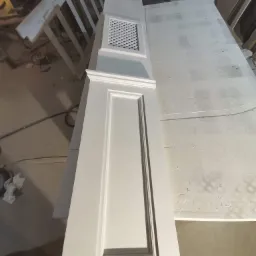 Biały, pomalowany element szafy z kratką wentylacyjną, leżący na stole w warsztacie stolarskim, widoczne inne elementy w tle.