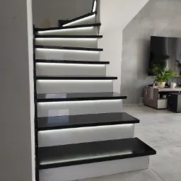 Nowoczesne schody z czarnymi stopniami i białymi podstopnicami, podświetlone ledami, widok z dołu, w tle salon z telewizorem.