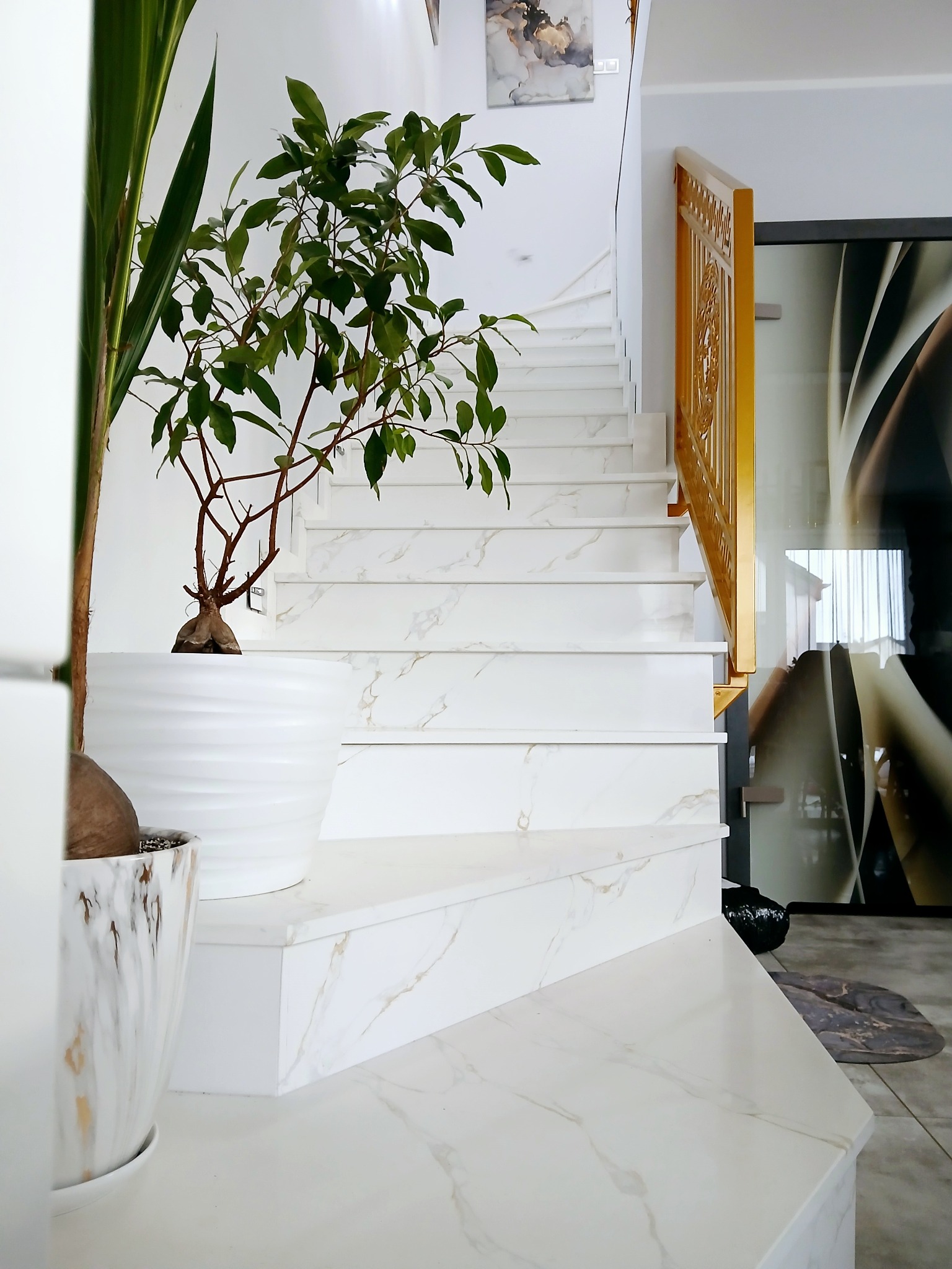 Białe schody z marmurowym wzorem, złota balustrada, rośliny doniczkowe w nowoczesnym wnętrzu.