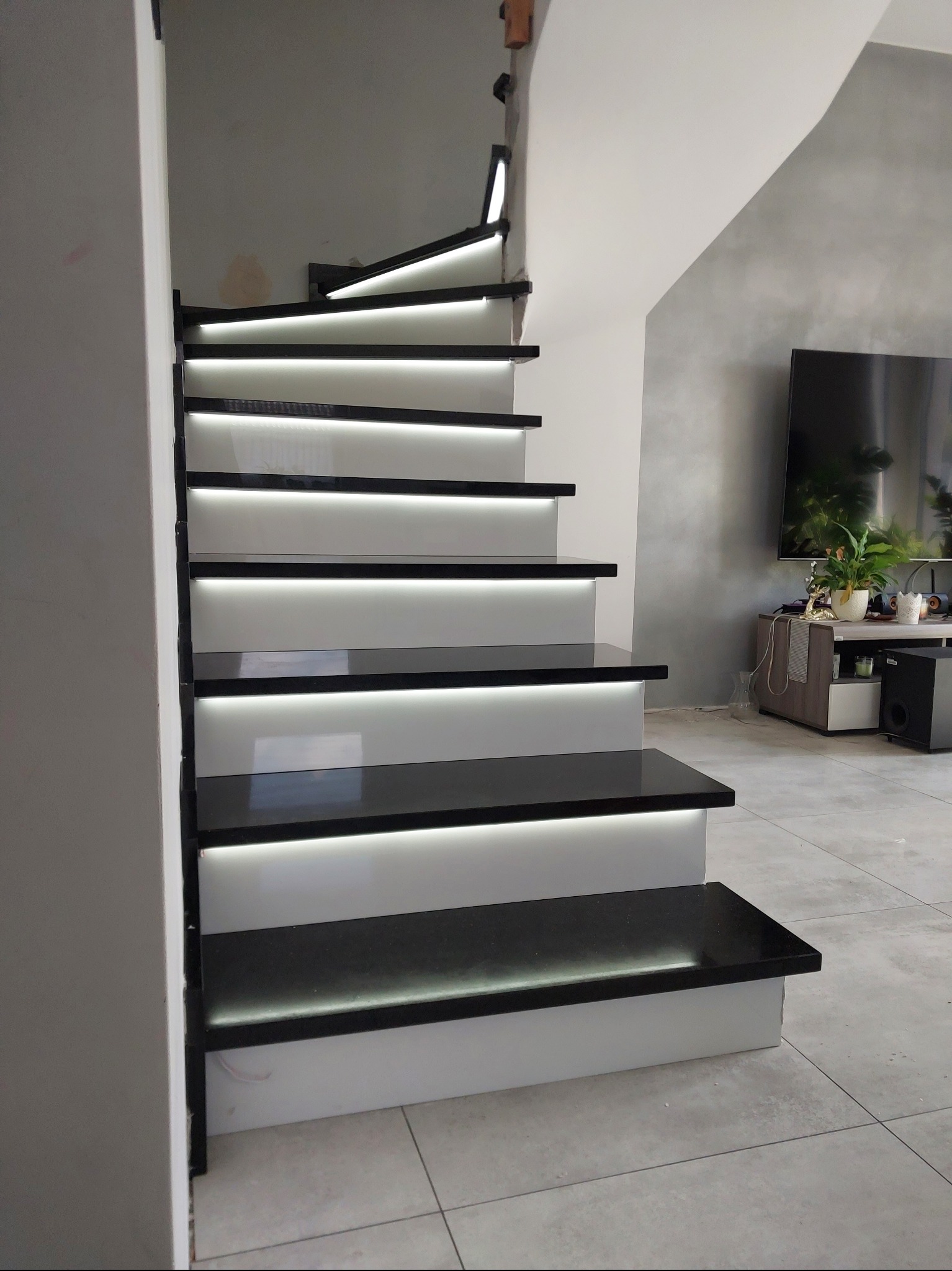 Nowoczesne schody z czarnymi stopniami i białymi podstopnicami, podświetlone ledami, widok z dołu, w tle salon z telewizorem.