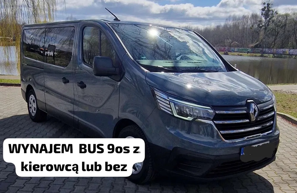 Szary bus Renault Trafic z napisem 'Wynajem bus 9os z kierowcą lub bez' stoi na parkingu z kostki brukowej, w tle zieleń i woda.