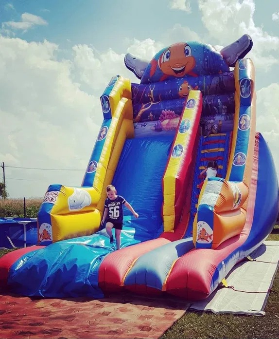 Dziecko zbiega z dmuchanej zjeżdżalni w kształcie ryby Nemo, na trawie, pod błękitnym niebem z chmurami. Zjeżdżalnia w kolorach niebieskim, żółtym i czerwonym.