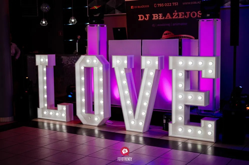 Podświetlany napis LOVE z żarówkami na parkiecie, w tle stanowisko DJ'a z fioletowym oświetleniem. Dekoracja sali weselnej.