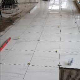 db BEAUTY - Układanie białych, marmurkowych płytek podłogowych w długim korytarzu, widoczne narzędzia i zaprawa, etap prac wykończeniowych.