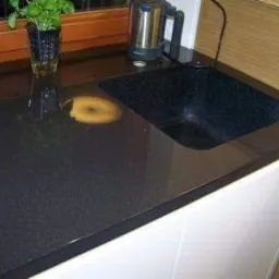 Czarny, błyszczący blat kuchenny z wbudowanym, ciemnym zlewem, uzupełniony o czajnik elektryczny i doniczkę z zieloną rośliną, widoczne białe szafki.