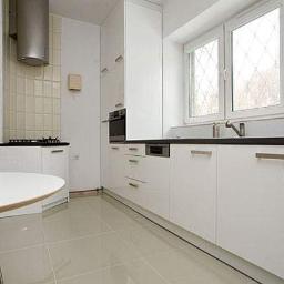 db BEAUTY - Jasna kuchnia z białymi, lakierowanymi szafkami, czarnym blatem i oknem z ozdobnymi szprosami; widoczny fragment okrągłego stołu z krzesłem.