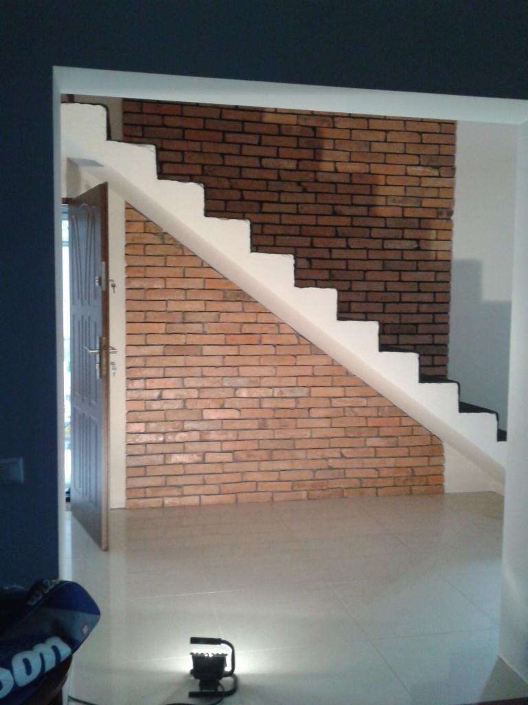 Klatka schodowa z ceglaną ścianą, białe stopnie, widoczna lampa robocza na podłodze.